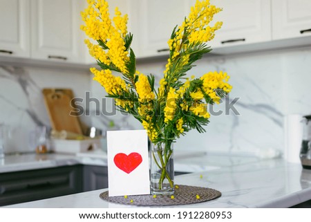 Mimosa Branch Postcard with a Heart on the background of a Gray and White Kitchen. Banner for March 8. Women's Day. Color 2021