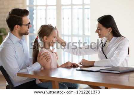 Caring young Caucasian female doctor cheer little girl child patient at hospital visit with dad. Attentive woman nurse or GP comfort caress small kid at consultation in clinic. Healthcare concept. Royalty-Free Stock Photo #1912281373