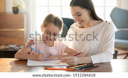 Smiling young Caucasian mother and little 7s cute daughter have fun drawing in album with pencils together. Happy mom and small girl child paint involved in funny learning activity. Hobby concept.