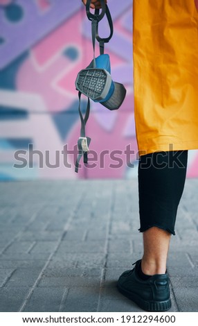 Cropped image of street artist with respirator mask in hand standing near the wall with his paintings. Street art concept