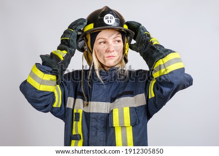 young brave woman in uniform of firefighter puts hardhat on her head and looking away on gray background