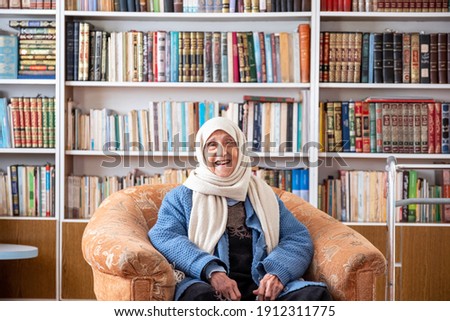 Arabic old woman sitting on couch in her home with a library background Royalty-Free Stock Photo #1912311775