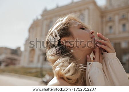 Curly blonde young woman in pearl jewelry smiles outside. Pretty attractive lady in stylish white blouse poses in city center.