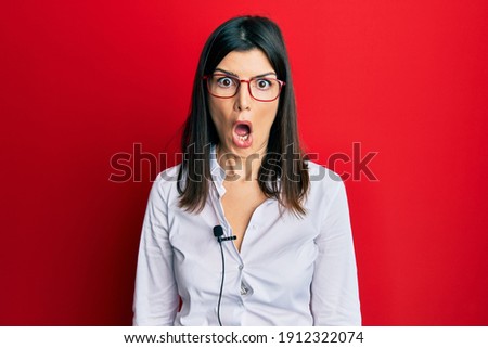 Young hispanic woman using lavalier microphone scared and amazed with open mouth for surprise, disbelief face 
