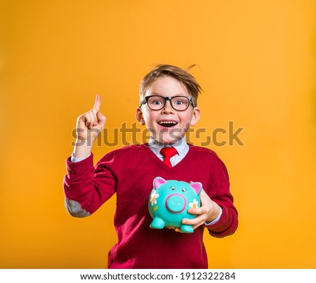Happy school boy with money pointing up isolated on yellow. Cool stylish businessman. How to be rich. Portrait of a cute nerdy boy carrying a piggy bank with his savings. Success, motivation, winner.