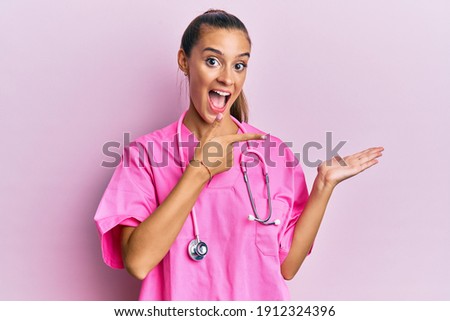 Young hispanic woman wearing doctor uniform and stethoscope amazed and smiling to the camera while presenting with hand and pointing with finger. 
