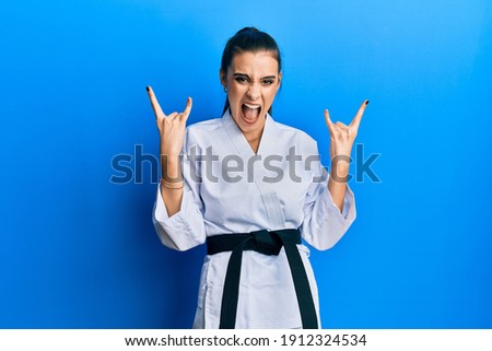 Beautiful brunette young woman wearing karate fighter uniform with black belt shouting with crazy expression doing rock symbol with hands up. music star. heavy concept. 