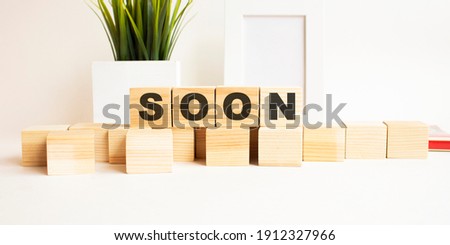 Wooden cubes with letters on a white table. The word is SOON. White background with photo frame, house plant.
