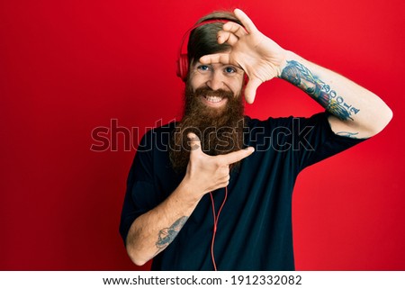 Redhead man with long beard listening to music using headphones smiling making frame with hands and fingers with happy face. creativity and photography concept. 