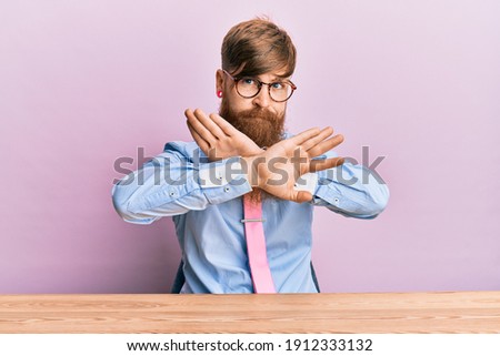 Young irish redhead man wearing business shirt and tie sitting on the table rejection expression crossing arms and palms doing negative sign, angry face 