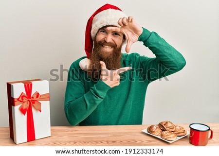 Young irish redhead man wearing christmas sitting on the table with gift smiling making frame with hands and fingers with happy face. creativity and photography concept. 