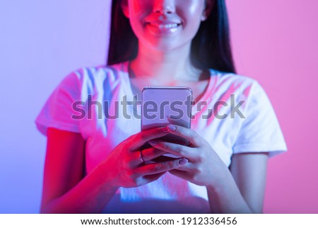 Chat on social networks, write blog, post online and surfing in internet. Cheerful asian zoomer female looks in glowing phone screen, typing message in neon, studio shot, cropped, close up, free space