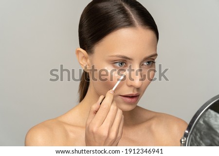 Woman with natural makeup applying concealer on flawless fresh skin, doing make up, looking at mirror. Girl after shower put corrector under eye area. Beauty face, skin care. Copy space, advertising. Royalty-Free Stock Photo #1912346941