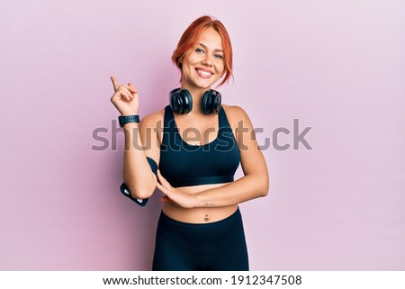 Young beautiful redhead woman wearing gym clothes and using headphones with a big smile on face, pointing with hand and finger to the side looking at the camera. 