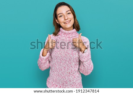 Young caucasian girl wearing wool winter sweater success sign doing positive gesture with hand, thumbs up smiling and happy. cheerful expression and winner gesture. 