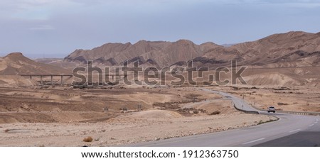 Asphalt road going through the Negev Desert near Big Crater in Israel. Chain of varicolored mountain ridges on the background. Dramatic, colorful and diverse desert landscape. Yellow sandstone hills.