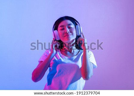 Stay alone at home during covid-19 and relax at spare time with music. Smiling calm millennial asian lady in modern headphones with closed eyes enjoying song in audio app, in neon, studio shot Royalty-Free Stock Photo #1912379704