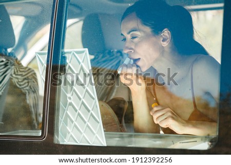 Attractive woman in bikini, looks at herself in a mirror while making up inside her camper van, road trip concept