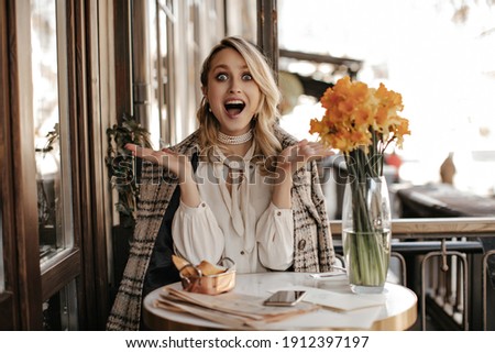Surprised young woman in white blouse and tweed checkered coat sits in cafe. Attractive girl looks shocked in restaurant.
