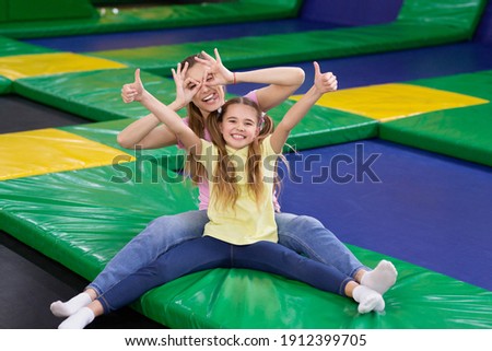 Playful silly mom with her teen daughter sitting at trampoline area of entertainment centre, showing thumbs up gesture. Mother and her child recommending to visit indoor kids playground Royalty-Free Stock Photo #1912399705