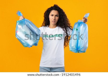 Displeased Female Volunteer Holding Two Plastic Garbage Bags Full Of Litter And Wasted Bottles Posing On Yellow Studio Background. Junk Sorting, Ecology Concept