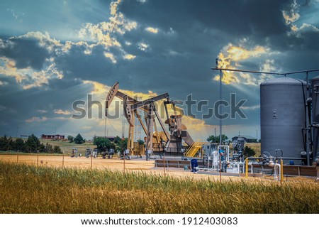 Two pump jacks and tanks and oil field equipment in fenced area in pasture under dramatic skies with houses on hill on horizon - summer