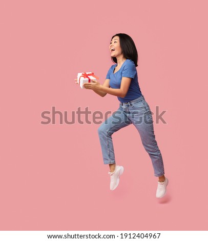 Full length vertical photo of jumping high excited asian lady holding present box looking away and giving gift to copy space isolated over pastel pink studio background. Greeting And Celebration