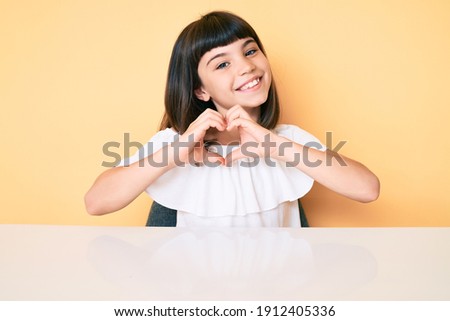 Young little girl with bang wearing casual clothes sitting on the table smiling in love doing heart symbol shape with hands. romantic concept.  Royalty-Free Stock Photo #1912405336