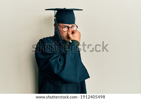 Young caucasian man wearing graduation cap and ceremony robe smelling something stinky and disgusting, intolerable smell, holding breath with fingers on nose. bad smell 