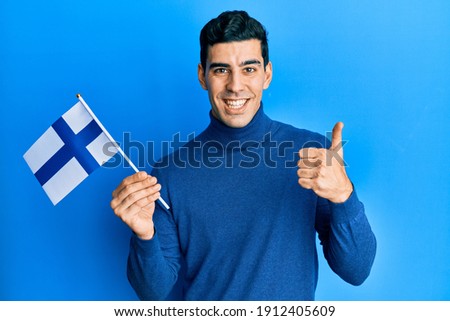 Handsome hispanic man holding finland flag smiling happy and positive, thumb up doing excellent and approval sign 