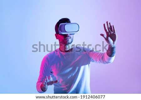 Amazing technology, online game, entertainment, study and virtual world in 3D simulation. Serious millennial man in vr glasses plays and tries to touch something, in neon, studio shot, free space Royalty-Free Stock Photo #1912406077