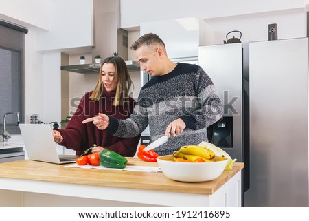 Stock photo of a young couple enjoying and preparing healthy meal in the kitchen and watching recipes on the laptop. Learning cooking and enjoying vegan meal