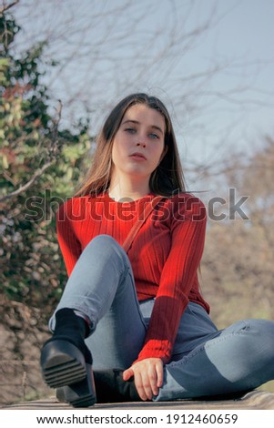 Close up picture of young thoughtful woman in red sweater and black high heels looking in front at the camera in a winter sunny day while sitting on a bench in side of a park.