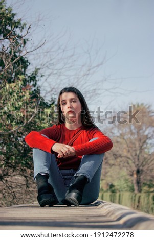 Open picture of young thoughtful woman in red sweater and black high heels looking in front at the camera in a winter sunny day while sitting on a bench in side of a park.