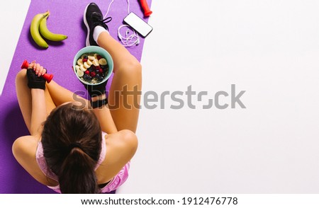 Sports and nutrition. Top photo of a girl with dumbbells and muesli in hand while sitting on the mat. White background and blank advertising space. Royalty-Free Stock Photo #1912476778