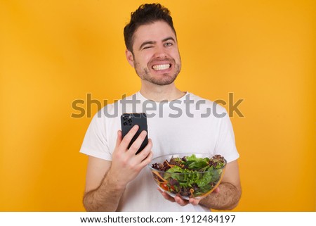 Pleased Young handsome Caucasian man holding a salad bowl against yellow background using self phone and looking and winking at the camera. Flirt and coquettish concept.