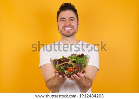 Positive glad Young handsome Caucasian man holding a salad bowl against yellow background says: wow how exciting it is, has amazed expression, shows something on blank space with open hand. 