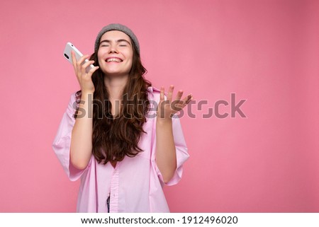 Positive attractive young brunette woman wearing stylish pink shirt and grey hat isolated over pink background holding in hand and using mobile phone communicating and recording voice message