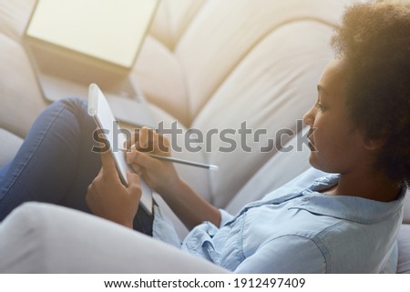 Close up shot of mixed race teen schoolgirl using pencil for making notes while doing homework, sitting on the couch at home