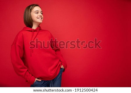 Photo shot of attractive happy positive smiling brunette female teenager wearing stylish red hoodie standing isolated over red background wall looking to the side