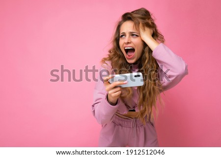 Photo of attractive crazy amazed surprised young woman wearing casual stylish clothes standing isolated over background with copy space holding and using mobile phone looking to the side and shouting