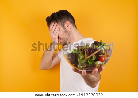 Young handsome Caucasian man holding a salad bowl against yellow background covers eyes with palm and doing stop gesture, tries to hide. Don't look at me, I don't want to see, feels ashamed or scared.