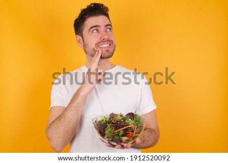 Young handsome Caucasian man holding a salad bowl against yellow background with thoughtful expression, looks to the camera, keeps hand near face, bitting a finger thinks about something pleasant.