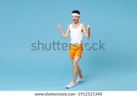 Full length portrait joyful young sporty fitness man with skinny body sportsman in white headband shirt shorts doing winner gesture isolated on blue background. Workout gym sport motivation concept Royalty-Free Stock Photo #1912521340