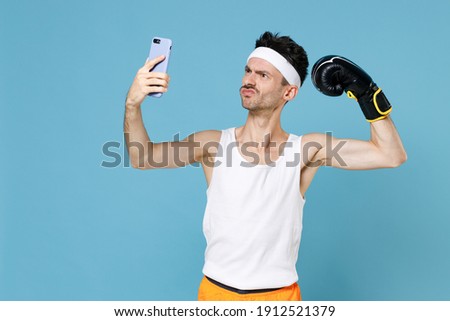 Confused man boxer with skinny body sportsman in headband shirt shorts boxing gloves doing selfie shot on mobile phone showing biceps muscles isolated on blue background. Workout gym sport concept