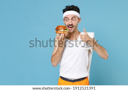 Amazed young sporty man with skinny body sportsman in headband shirt towel hold american classic fast food burger showing thumb up isolated on blue background. Workout gym sport motivation concept