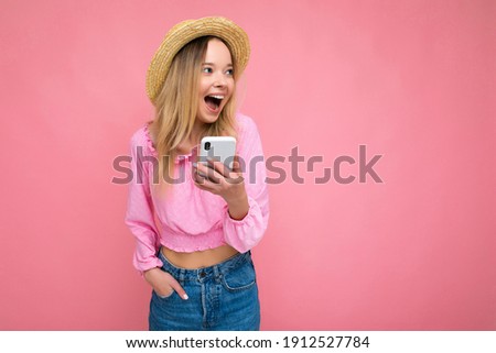 Attractive amazed young blonde woman smiling and having fun standing isolated over pink wall keeping hand in pocket wearing pink blouse and summer hat using mobile phone looking to the side