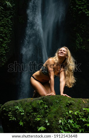 Young Caucasian woman with long blond hair sitting on the rock near the waterfall. Happy smiling woman. Travel lifestyle. Woman wearing bikini. Leke Leke waterfall, Bali, Indonesia