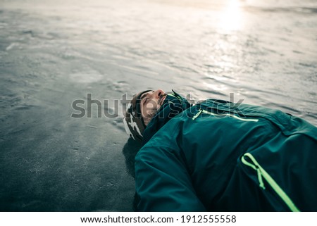 Portrait of a beautiful young European man in green jacket lying on a blue ice-frozen lake with snow and cracks in winter. Reflection, drawing of nature, clouds, blue water, abstraction, cold, travel