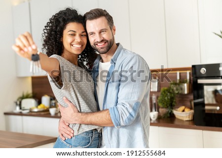 Young interracial married couple homeowners smiling, showing keys from a new apartment, hugging and looking at the camera, standing in the kitchen and celebrating moving in a new home, family concept Royalty-Free Stock Photo #1912555684